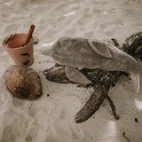 The Senger large organic cotton stuffed dolphin toy, posed on a piece of driftwood on a sandy beach