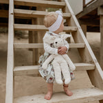 A child holding onto a Senger large floppy white rabbit with both hands. The child is sitting on wooden steps on a beach. 