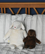 A Senger large white sheep and small brown sheep soft toy in seated position next to each other, leaning against pillows and a wooden headboard. These sheep are part of a wide range of soft toys made from organic and natural materials stocked at Babipur. 