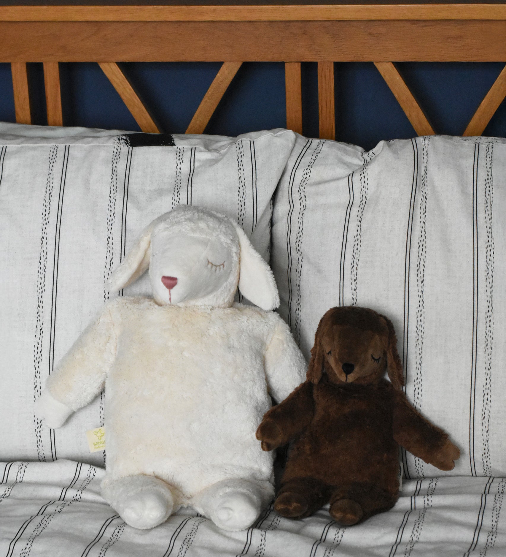 A Senger large white sheep and small brown sheep soft toy in seated position next to each other, leaning against pillows and a wooden headboard. These sheep are part of a wide range of soft toys made from organic and natural materials stocked at Babipur. 