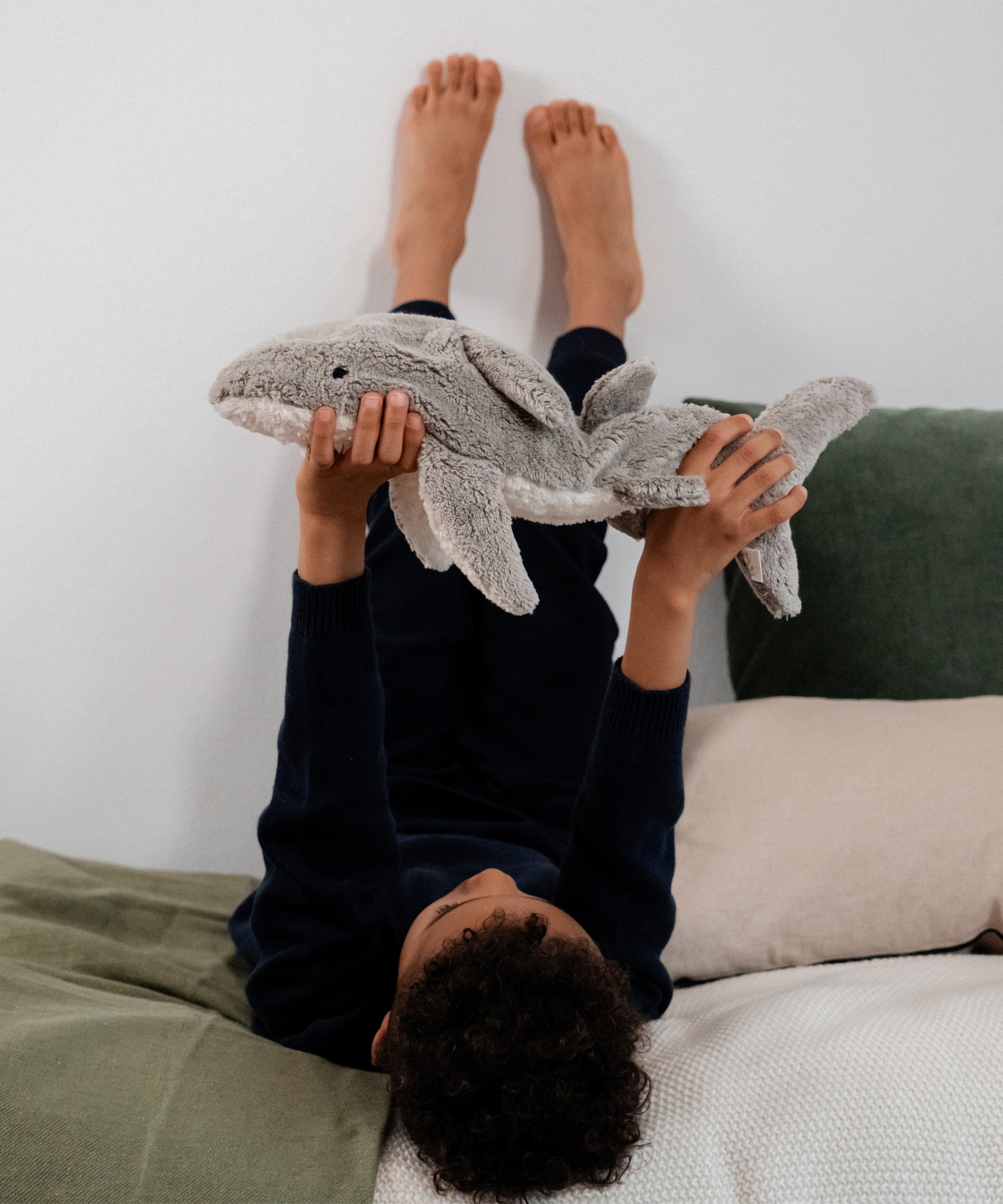 Child lying on a bed holding the Senger Naturwelt Small Cuddly Shark, organic heatable soft toy with grape seed cushion.