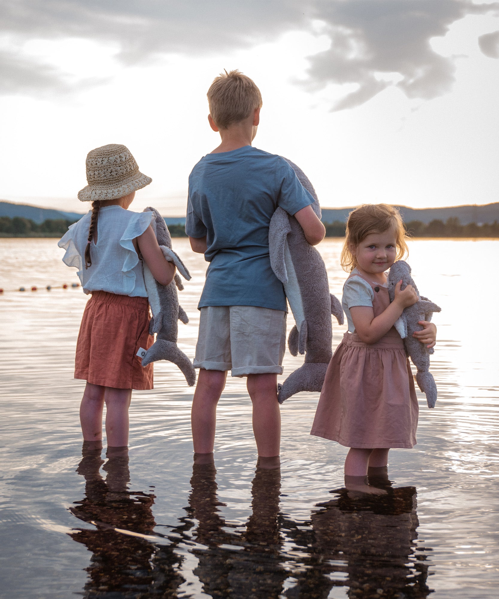 Children standing in shallow water holding the Senger Naturwelt Cuddly Shark, organic GOTS cotton heatable soft toy.