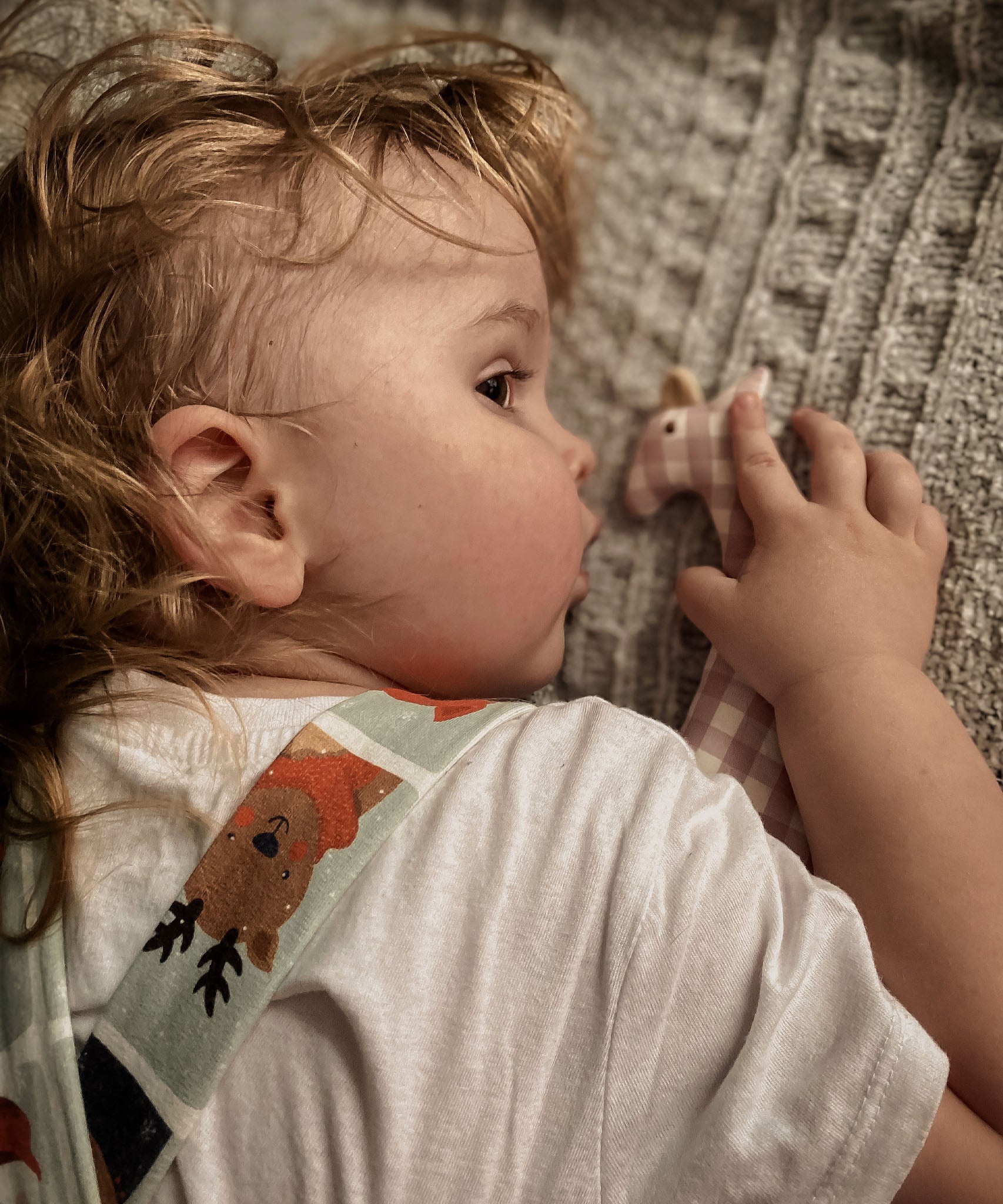 A child laying down next to a Senger organic cotton pink and white check giraffe soft grasping toy, available at Babipur.