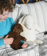 A Senger small brown cuddly sheep soft toy in placed on a child's lap in bed. The child is holding one of the sheep's hands. A large white cuddly sheep can be seen next to the child. These sheep are part of a wide range of soft toys made from organic and natural materials stocked at Babipur. 
