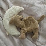 A small Senger brown camel cuddly toy lying flat next to a moon cushion on a blanket