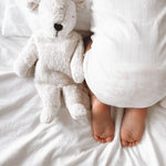 A closeup of a sleeping baby on white sheets, with a small Senger cuddly polar bear teddy