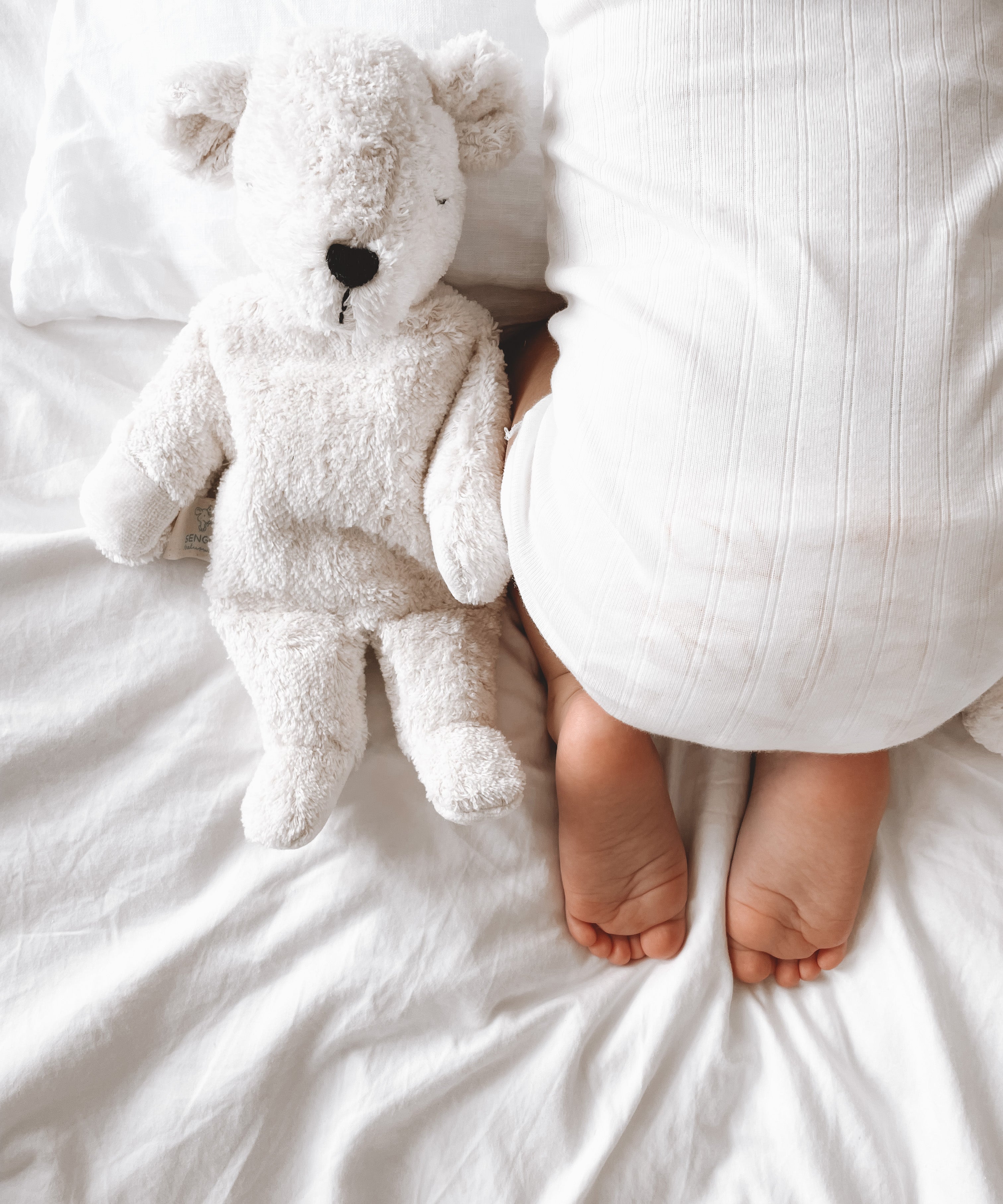 A closeup of a sleeping baby on white sheets, with a small Senger cuddly polar bear teddy