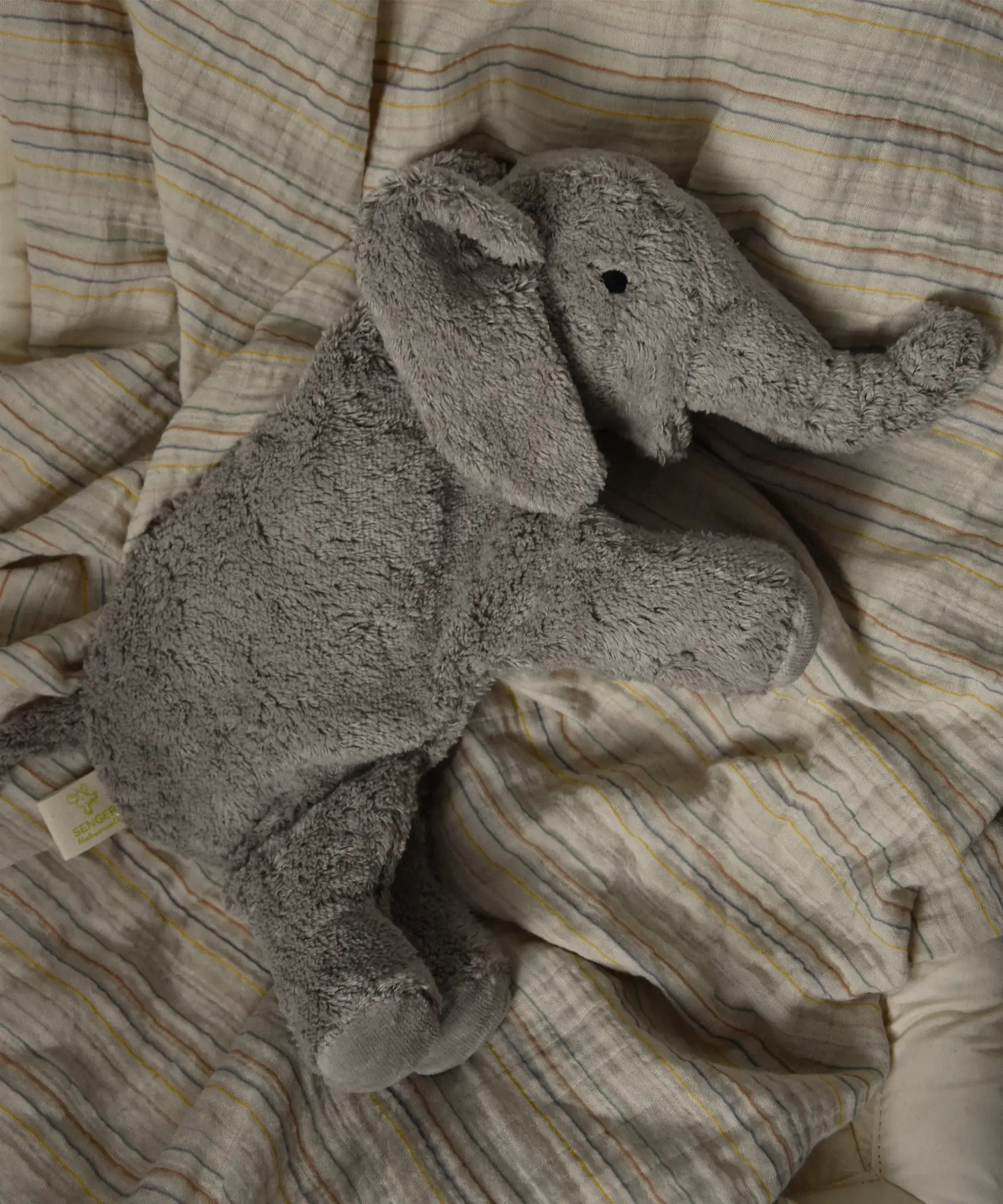 Small Senger elephant cuddly toy lying flat on a striped blanket