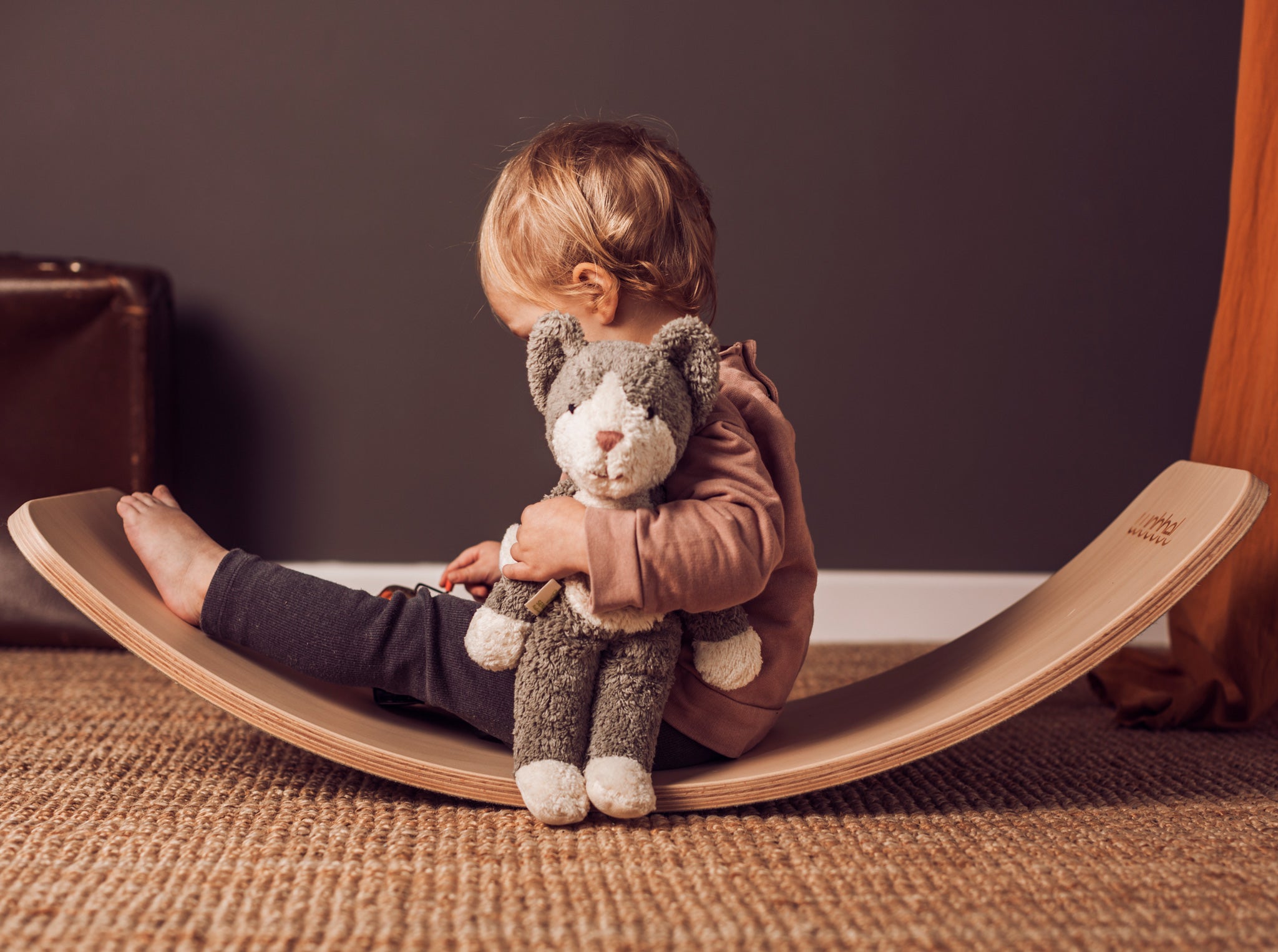 A child sitting on a Wobbel board holding a Senger Small Floppy Cat in one arm.