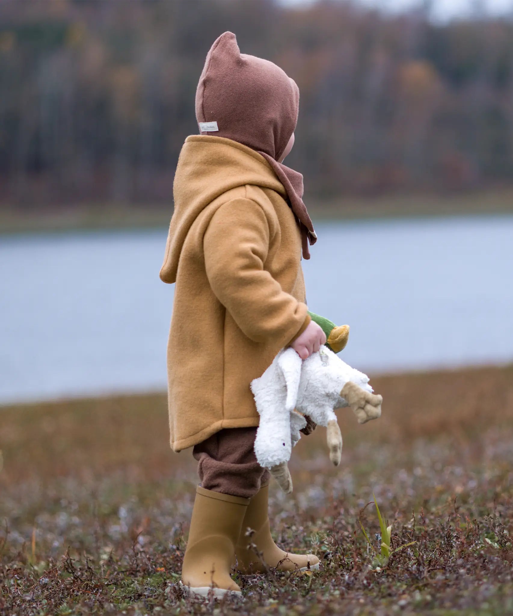 Child with a Senge small white goose cuddly toy in their hand. Standing on a grassy surface and a river in the background