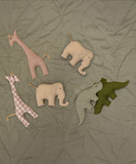 A group of soft grasping toys from Senger available at Babipur on a soft green blanket showing the pink giraffe, beige/cream elephant and the green/plaid stegosaurus soft toys.