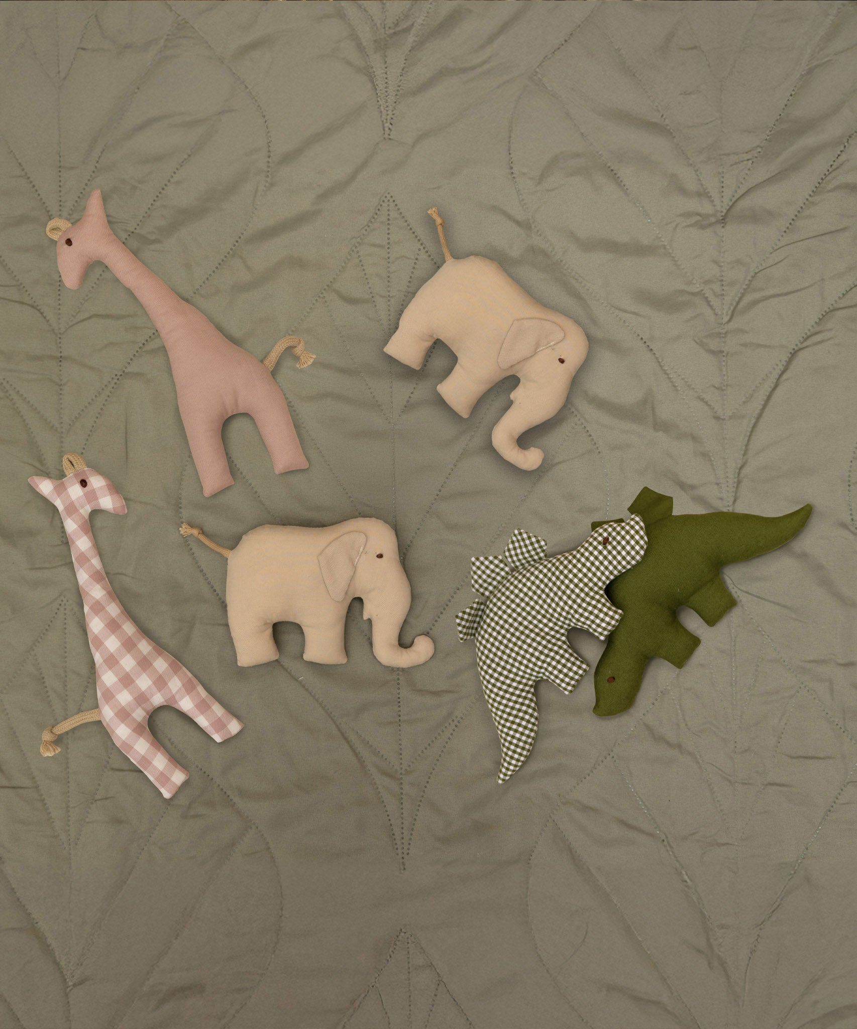 A group of soft grasping toys from Senger available at Babipur on a soft green blanket showing the pink giraffe, beige/cream elephant and the green/plaid stegosaurus soft toys.