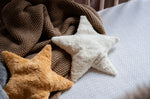 Two Senger star shaped warming cushions placed on a bed on a brown coloured blanket. One is a light yellow, off white colour and the other is a bright yellow colour. 