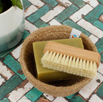 A Slice of Green beech wood nail scrubbing brush on top of a soap bar in a woven rope bowl