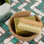 A Slice of Green beech wood nail scrubbing brush on top of a soap bar in a woven rope bowl