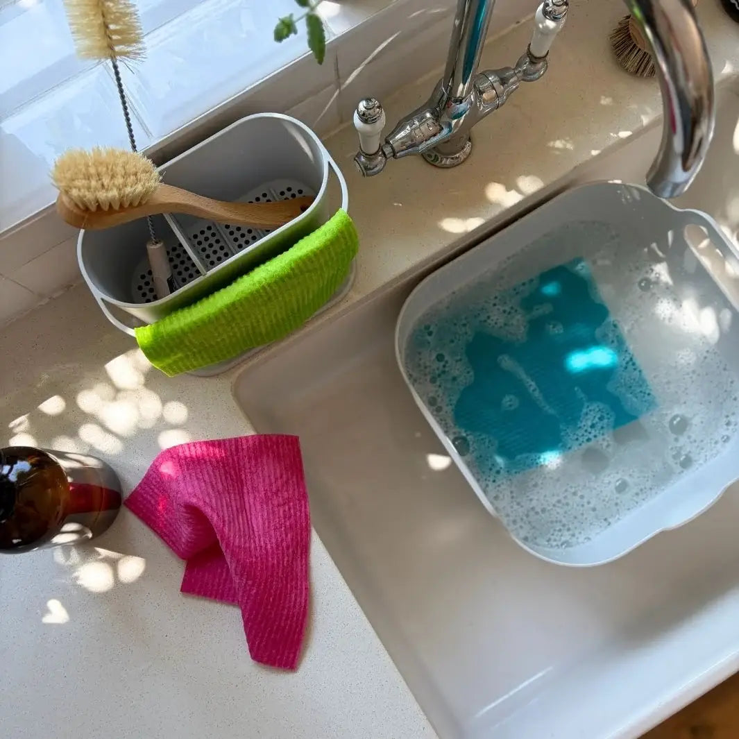 Bright pink, blue, and green Swedish sponge cloths being used and washed around a kitchen sink in a plastic-free kitchen.