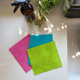 Three natural material reusable Swedish sponge cloths in bright pink, blue, and green beside a kitchen sink.