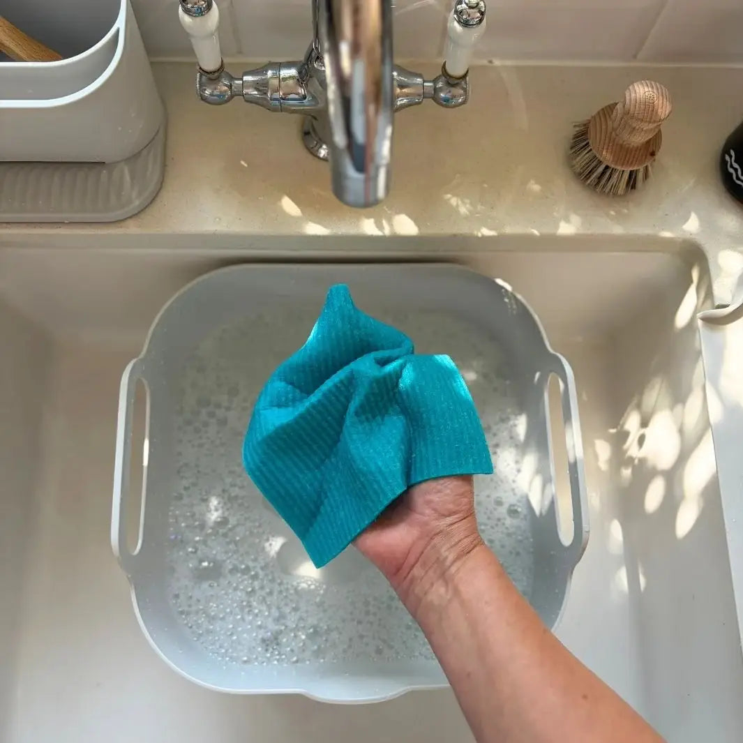 A Slice of Green Swedish sponge cloth made from natural materials being rinsed by hand over soapy water in a kitchen sink.