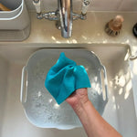 A Slice of Green Swedish sponge cloth made from natural materials being rinsed by hand over soapy water in a kitchen sink.