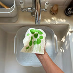 A reusable Swedish sponge cloth with a green botanical print being hand-washed over a kitchen sink in a plastic-free home.