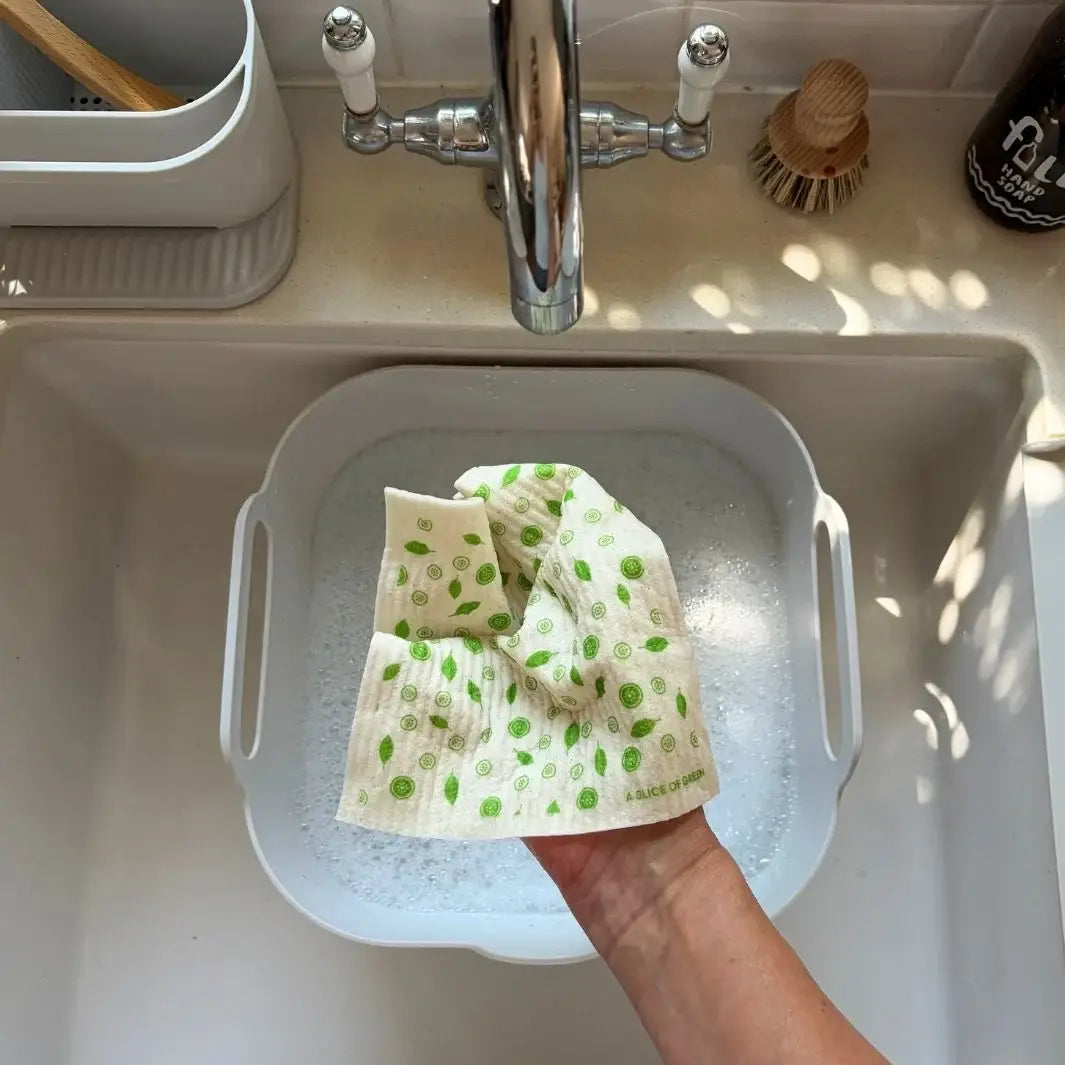 A reusable printed Swedish sponge cloth being hand-washed over a bowl of soapy water in a plastic-free kitchen sink.