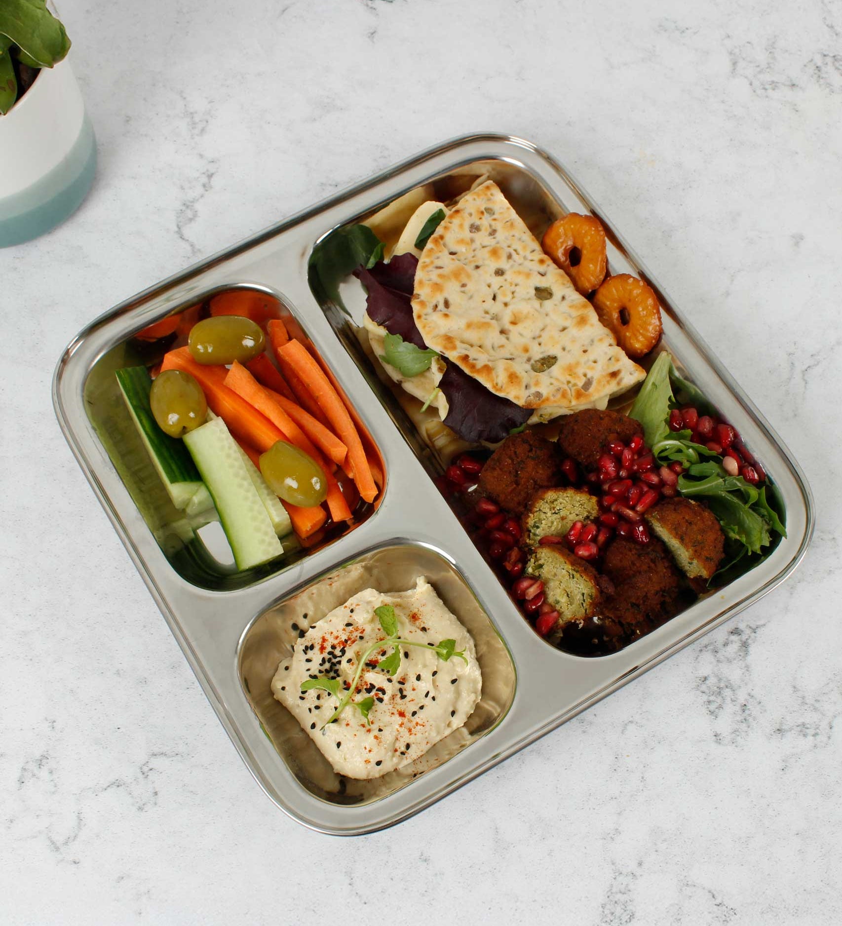 A Slice Of Green Stainless Steel Divided Plate. The plate has hummus in the smallest section, carrot, cucumber and olives in the middle section and flatbread, salad and falafels in the biggest section. This stainless steel plate is suitable for snacks and meals and perfect for children and adults. 