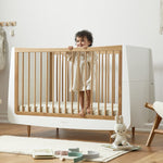 A SnuzKot Cot bed in oak pictured in a nursery. A child with curly hair can be seen standing up in the cot holding onto the side