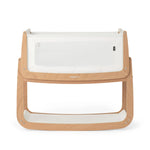The SnuzPod5 bedside baby crib with a natural, unpainted wooden frame, white mesh sides, and a white mattress. 
