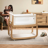 The natural wooden SnuzPod5 bedside crib with wheels, that has been wheeled into the living room whilst a mother holds her baby. 