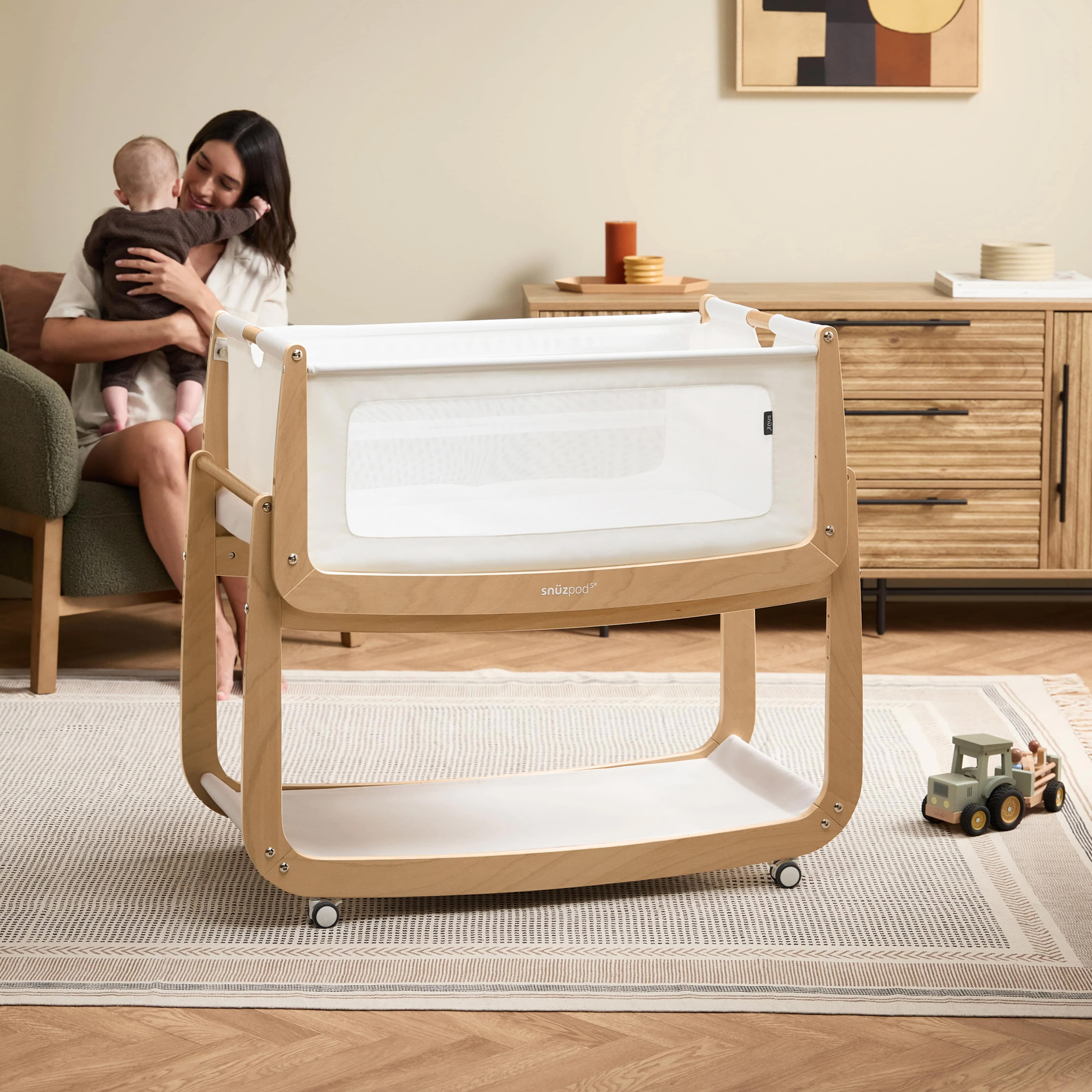 The natural wooden SnuzPod5 bedside crib with wheels, that has been wheeled into the living room whilst a mother holds her baby. 