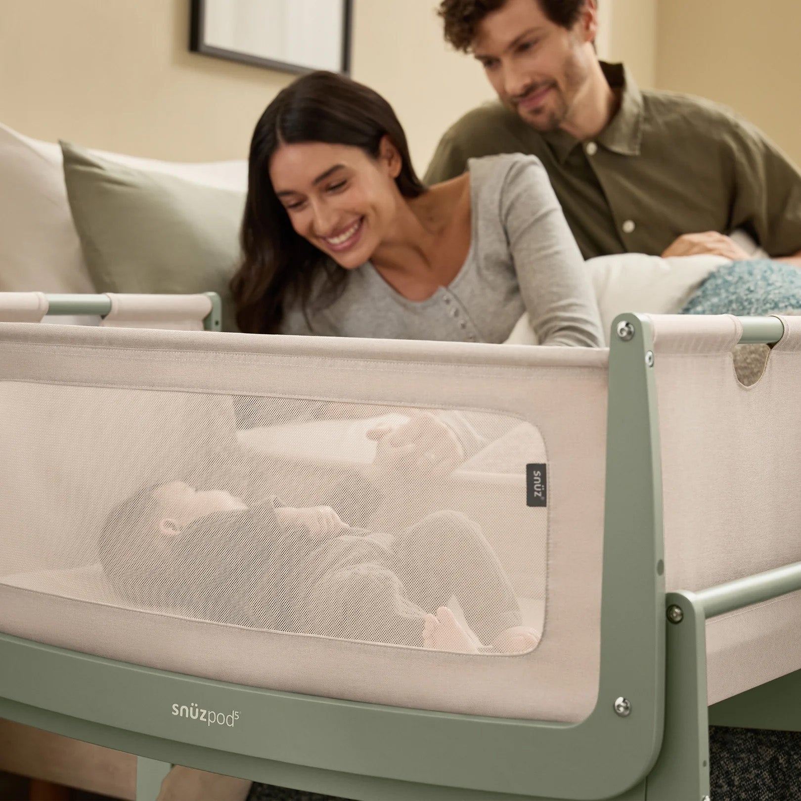 Two parents loving view their baby in the SnuzPod5 bedside crib, watching the baby from the comfort of their own bed.