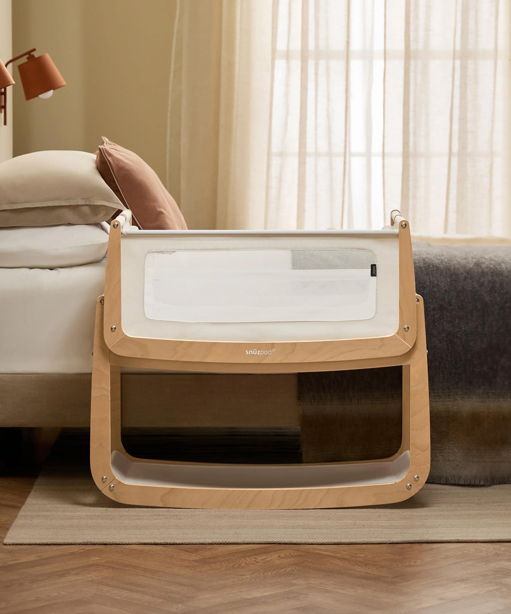 The SnuzPod 5 bedside baby crib with a lovely natural wooden frame, next to an adult's bed in a beautifully decorated bedroom.