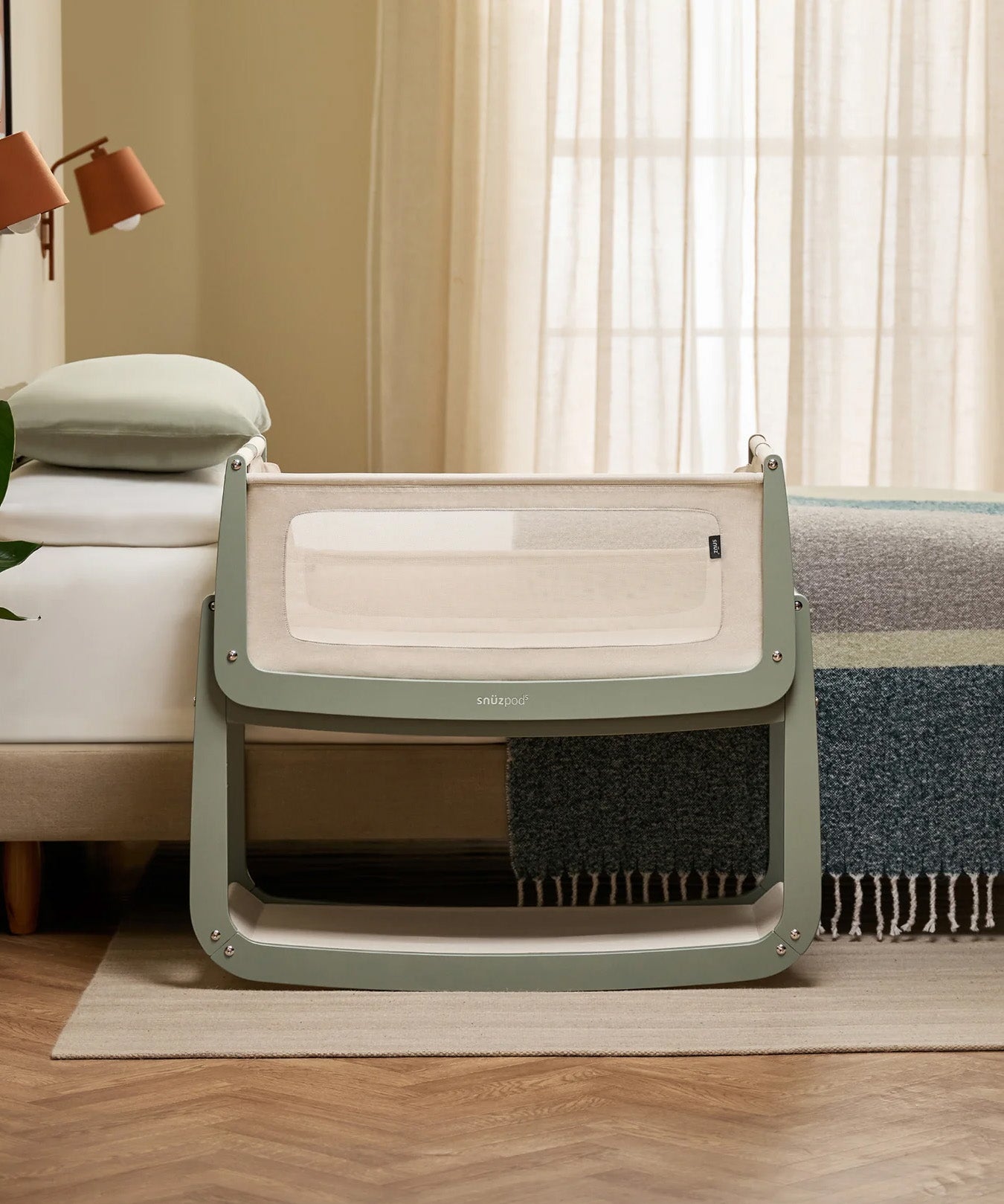 The SnuzPod 5 bedside baby crib with a lovely sage green wooden frame, next to an adult's bed in a beautifully decorated bedroom.