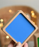 An adult's hand holding up the bottom blue piece from the Bajo Square Pyramid Sensory Wooden Stacking Toy. The set contains 5 wooden square and rectangular shaped blocks with a different coloured bases. Each blocks make a different sound when shaken. These heirloom quality toys by Bajo are part of a wide range of wooden toys available here at Babipur.  