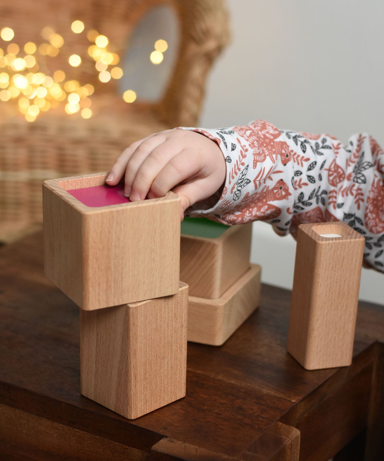 A close up of a child playing with the Bajo Square Pyramid Sensory Wooden Stacking Toy. The child is reaching for the pink cube block and placing it on the stack. The set contains 5 wooden square and rectangular shaped blocks with a different coloured bases. Each blocks make a different sound when shaken. These heirloom quality toys by Bajo are part of a wide range of wooden toys available here at Babipur. 