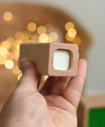 An adult's hand holding up the top piece from the Bajo Square Pyramid Sensory Wooden Stacking Toy. The set contains 5 wooden square and rectangular shaped blocks with a different coloured bases. Each blocks make a different sound when shaken. These heirloom quality toys by Bajo are part of a wide range of wooden toys available here at Babipur.  