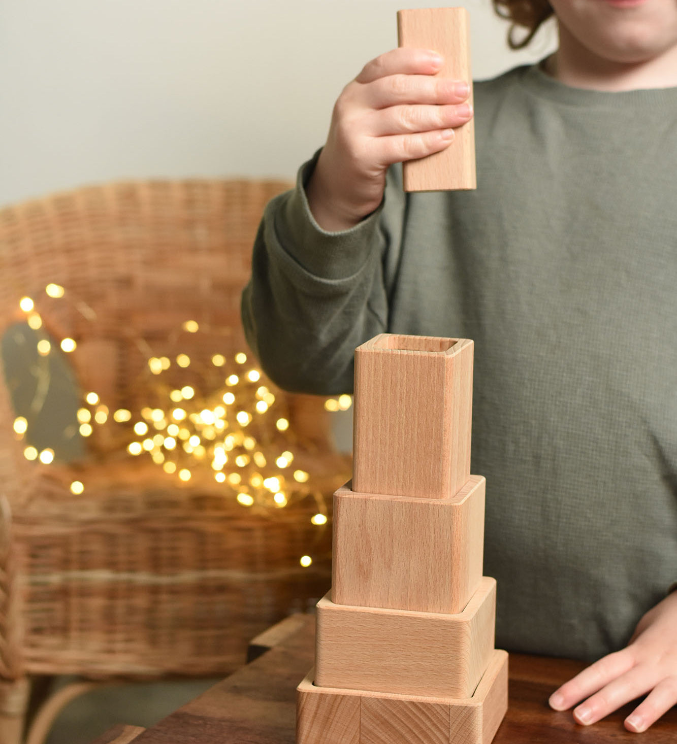 A close up of a child playing with the Bajo Square Pyramid Sensory Wooden Stacking Toy. The child has the tallest piece in their hand and they are about to place it on top of the stack. The set contains 5 wooden square and rectangular shaped blocks with a different coloured bases. Each blocks make a different sound when shaken. These heirloom quality toys by Bajo are part of a wide range of wooden toys available here at Babipur. 