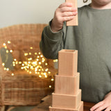 A close up of a child playing with the Bajo Square Pyramid Sensory Wooden Stacking Toy. The child has the tallest piece in their hand and they are about to place it on top of the stack. The set contains 5 wooden square and rectangular shaped blocks with a different coloured bases. Each blocks make a different sound when shaken. These heirloom quality toys by Bajo are part of a wide range of wooden toys available here at Babipur. 
