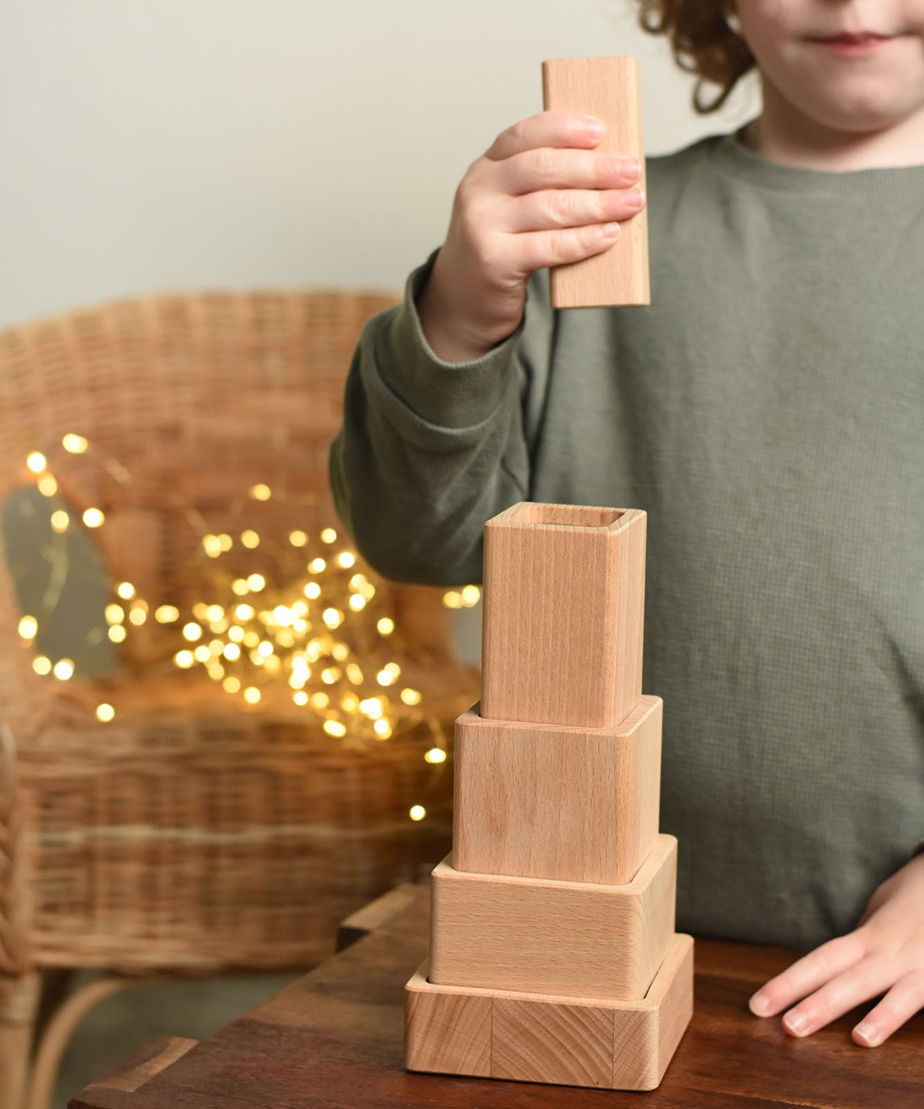 A close up of a child playing with the Bajo Square Pyramid Sensory Wooden Stacking Toy. The child has the tallest piece in their hand and they are about to place it on top of the stack. The set contains 5 wooden square and rectangular shaped blocks with a different coloured bases. Each blocks make a different sound when shaken. These heirloom quality toys by Bajo are part of a wide range of wooden toys available here at Babipur. 