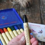 Black Stockmar wax crayon in an adult's hand. The metal storage tin can be seen in the background.