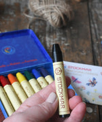 Black Stockmar wax crayon in an adult's hand. The metal storage tin can be seen in the background.