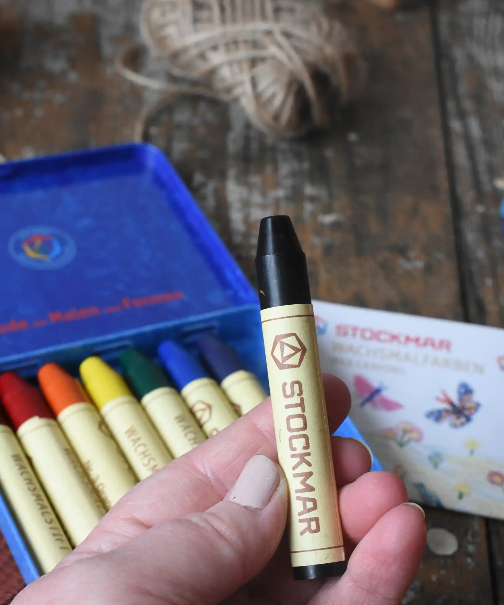 Black Stockmar wax crayon in an adult's hand. The metal storage tin can be seen in the background.