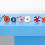 The 8 flowers from the Studio Roof Cardboard Bouquet in a  Klimt's Garden design placed on a blue surface all lined up. A DIY Cardboard craft kit that makes up 8 3D flowers. This 3D pop out DIY kit is part of Studio Roof's artist flowers collection which is available at Babipur. 