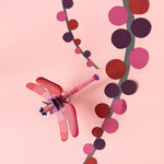 Studio Roof Biodegradable recyclable cardboard ruby dragonfly on a light pink/peach background with paper pink and red plants/flowers