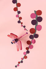 Studio Roof Biodegradable recyclable cardboard ruby dragonfly on a light pink/peach background with paper pink and red plants/flowers