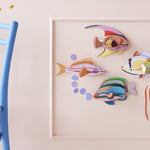 Studio Roof DIY craft fish wall decorations on a light pink wall next to a blue chair and glass vase