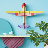 Studio Roof DIY card Plane wall decoration on a blue wall above a yellow shelf and a potted plant