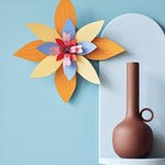 Studio Roof orange and blue craft flower hanging on a blue wall next to a light blue shelf with a brown candle holder