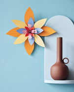 Studio Roof orange and blue craft flower hanging on a blue wall next to a light blue shelf with a brown candle holder