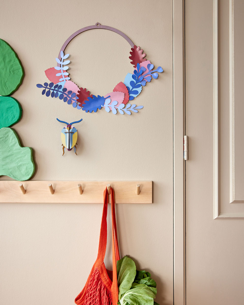 Studio roof pop out botanical wall decoration hanging on a beige wall above some wooden hangers and green leaves